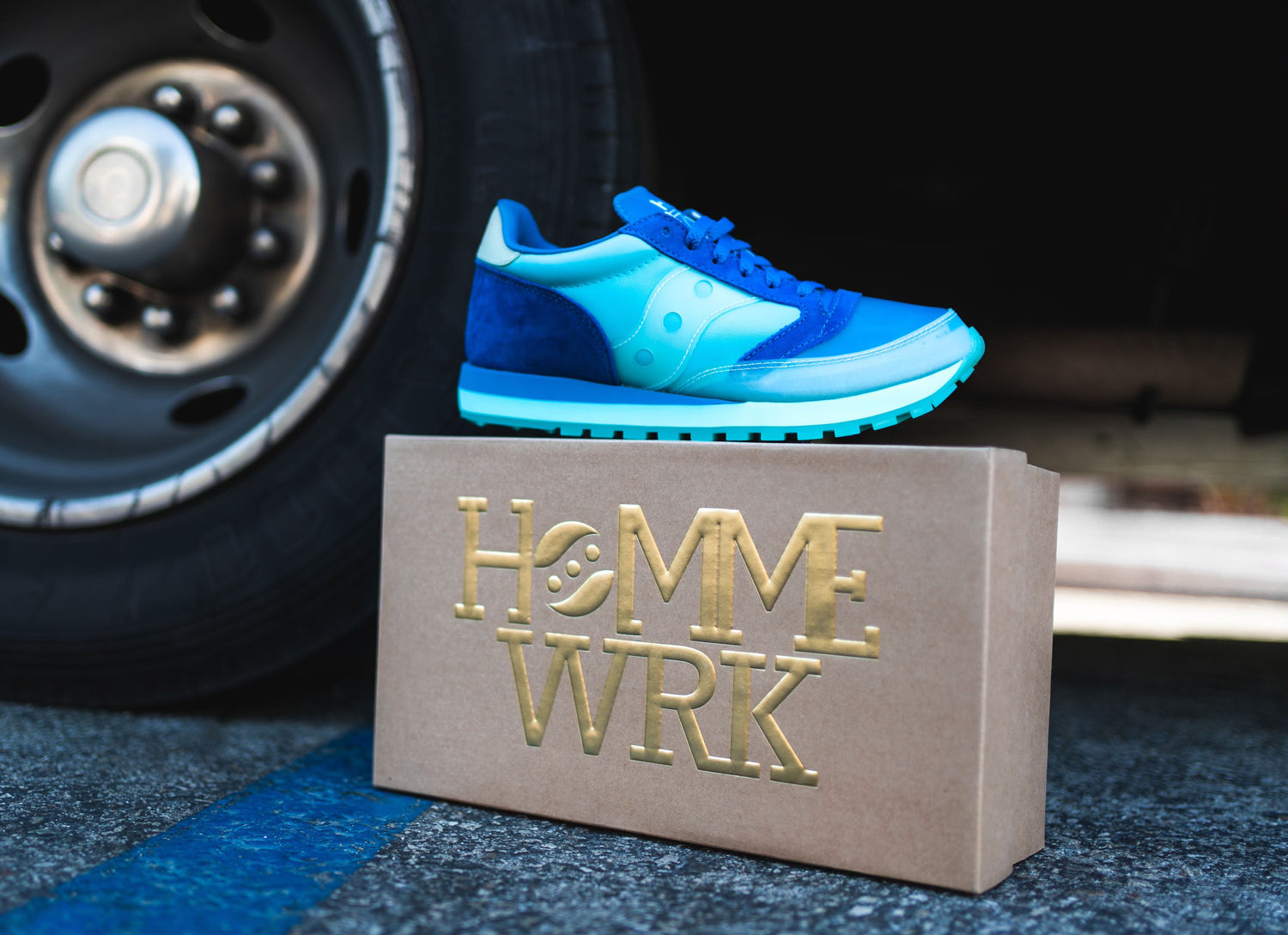 A pair of blue sneakers on top of a brown hommewrk shoe box in front of a cars wheel
